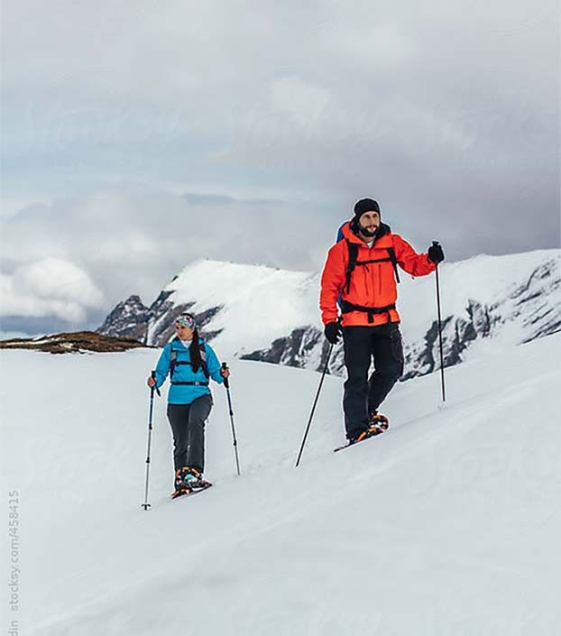 Man in orange coat and woman in teal coat snowshoeing on a mountain peak