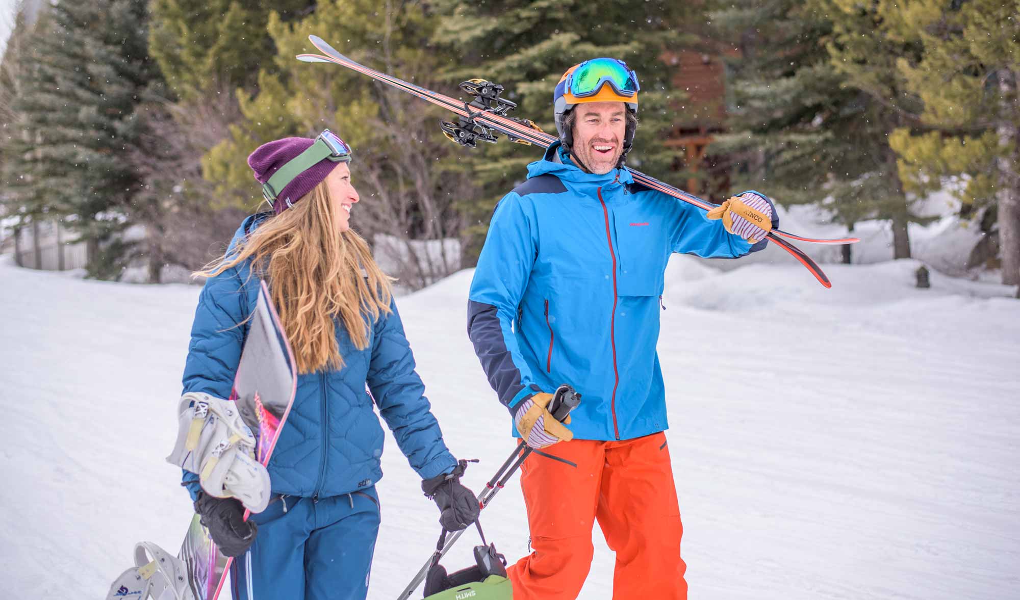 Man in blue coat and orange ski pants carrying skis and woman with blue coat and ski pants carrying snowboard