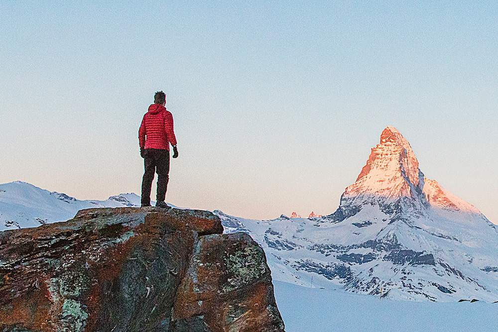 Man in red jacket standing on mountain rock looking at jagged peak with sun shining on it
