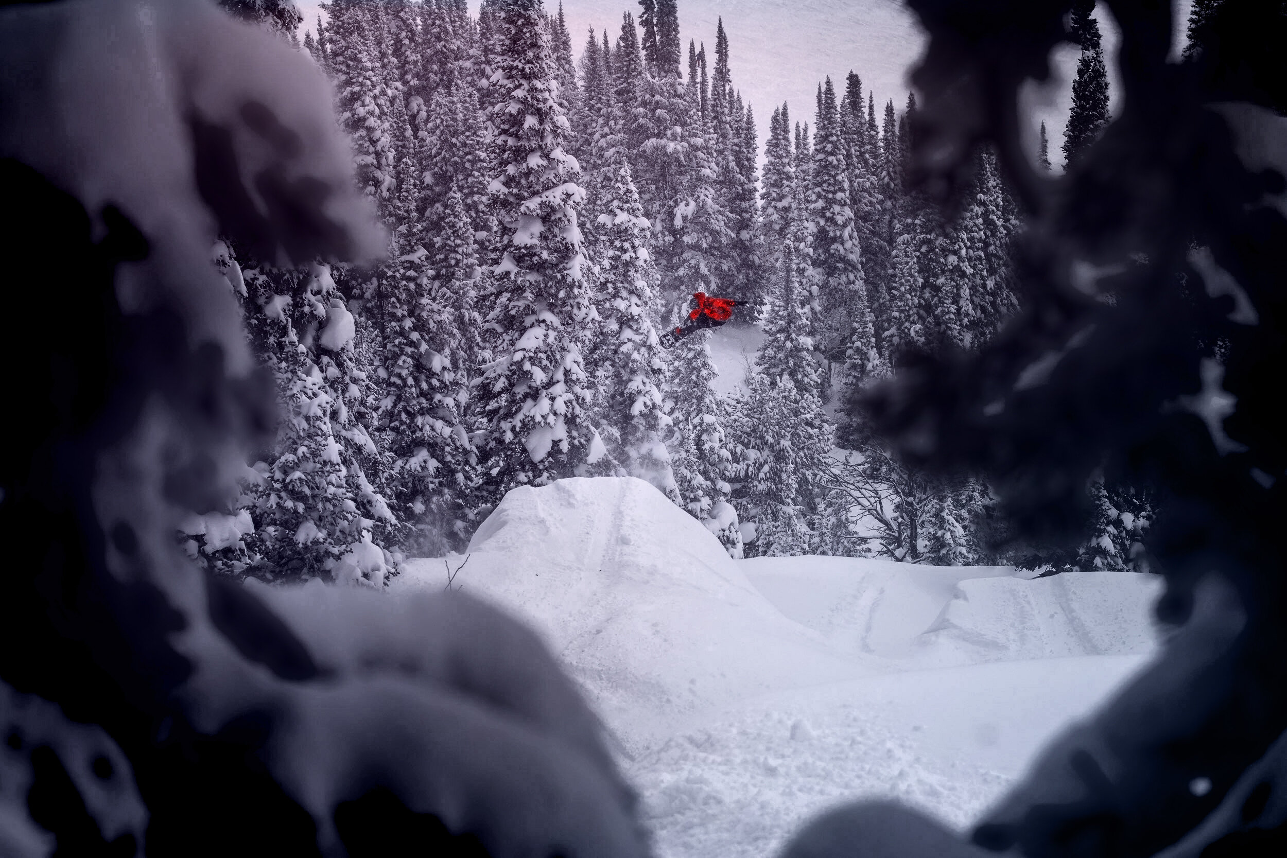 Snowboarder in red airborne after making jump surrounded by snow covered pine trees